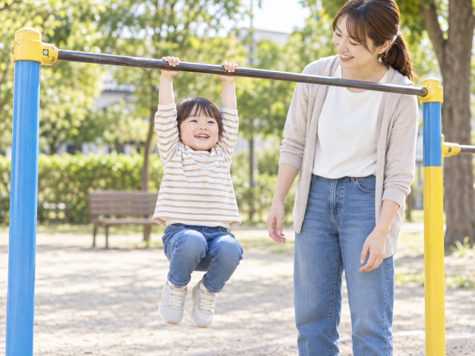 公園の鉄棒でぶら下がりを楽しむ幼児と見守る保護者